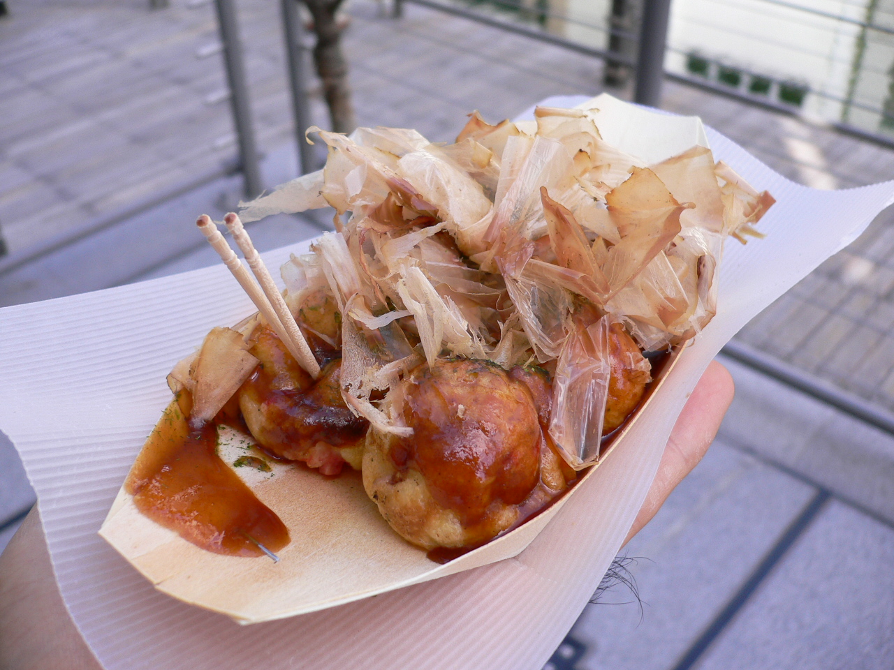 A warm food boat of fresh takoyaki sits atop a table drizzled in BBQ sauce and thin tuna flakes