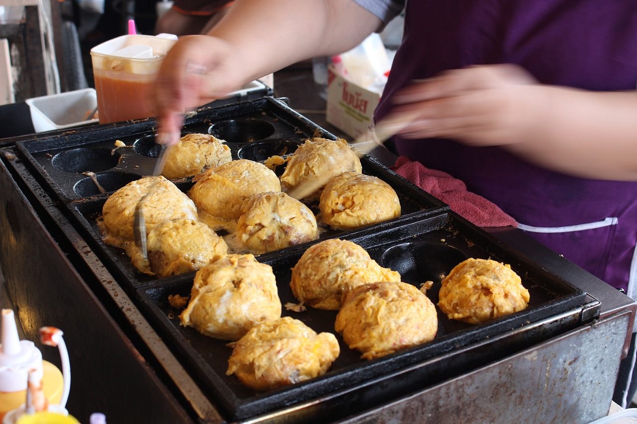 A street vendor diligently flips large doughy takoyaki balls as they cook in a special black takoyaki grill