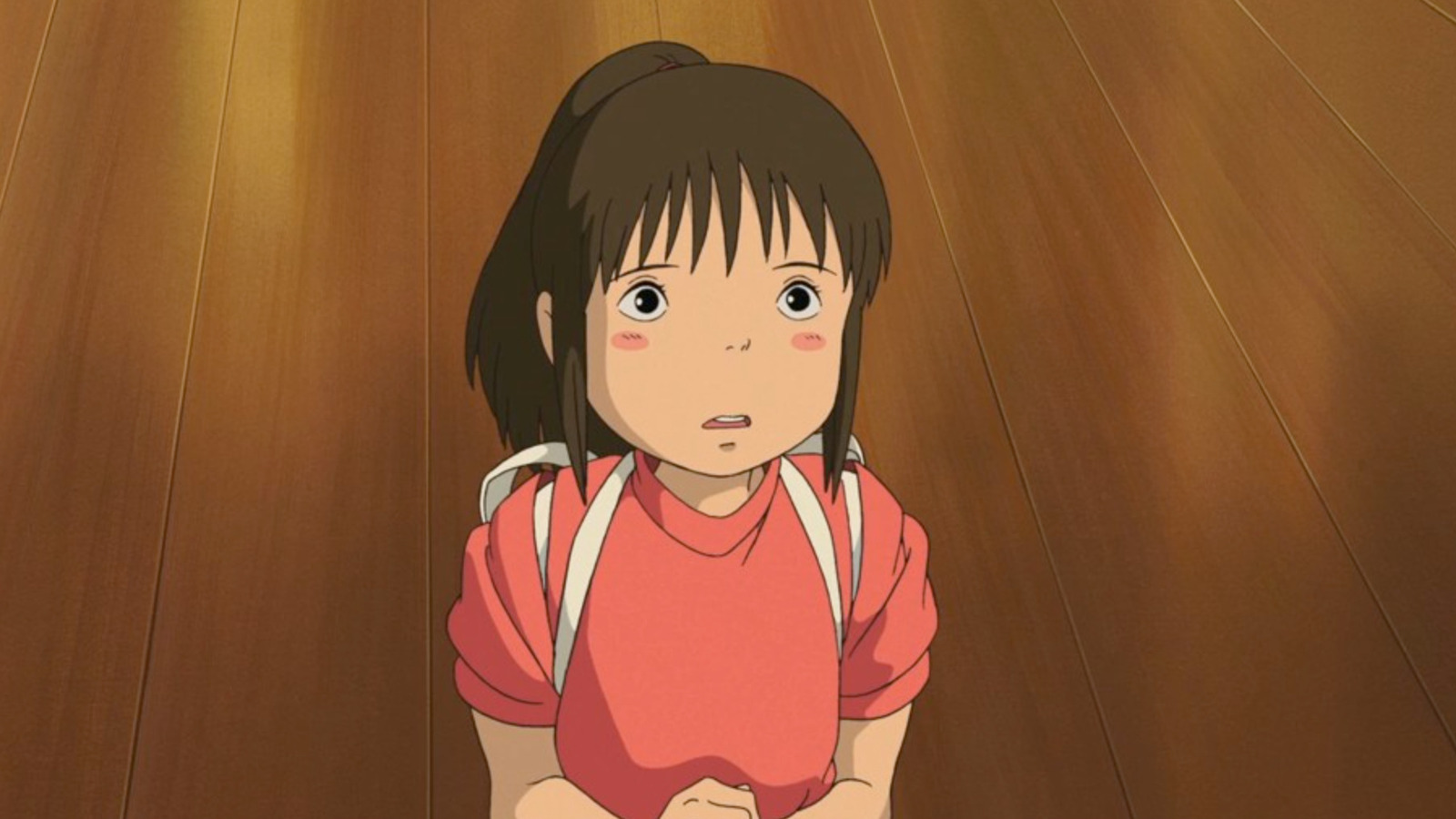 A young girl with her hair tied back looks up with uncertainty while standing in a soft red uniform