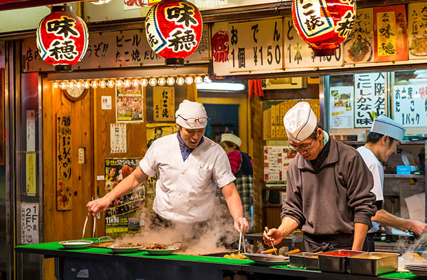 Two men dilegently flip little takoyaki balls as they book to perfection inside their Takoyaki street stand.