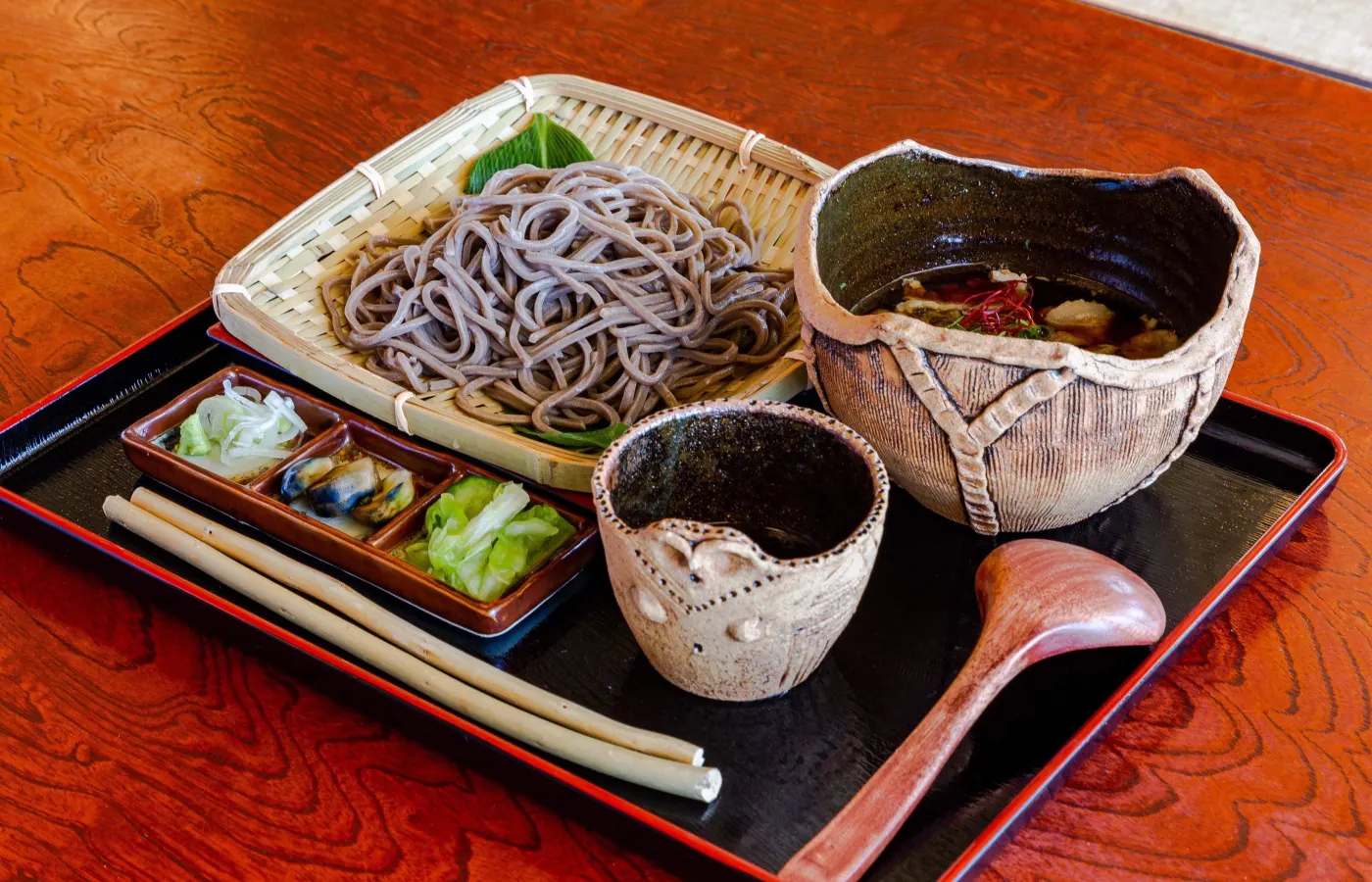 A slightly bowled plate of noodles with a side of soup, dipping sauce and pickled delights